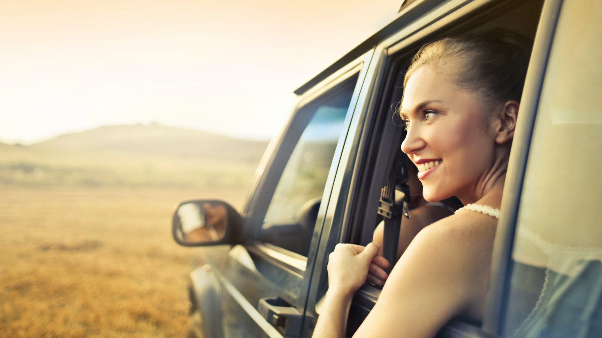 A person smiling looking out the window of a car in a rural landscape