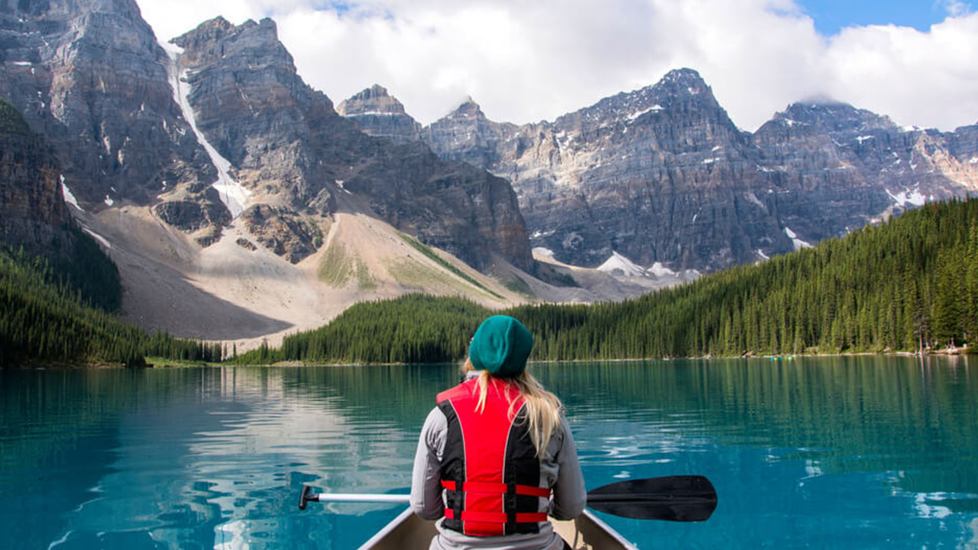 Person canoeing in clear blue waters looking at the dense forest and mountains in the distance