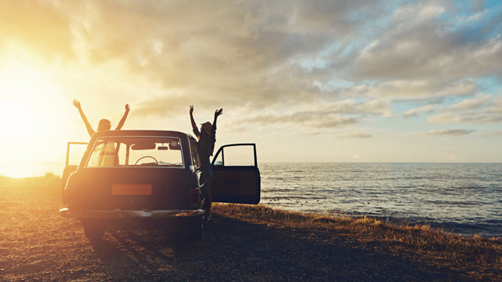 Two people cheering beside a car overlooking a beautiful sunset by a lake